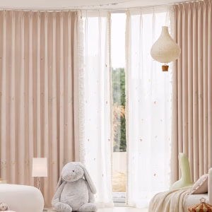 Zachtroze gordijnen met eenhoorns en maantjes, witte vitrage en een opgebonden rechtergordijn in een kinderkamer.