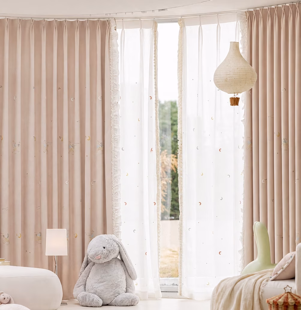 Zachtroze gordijnen met eenhoorns en maantjes, witte vitrage en een opgebonden rechtergordijn in een kinderkamer.